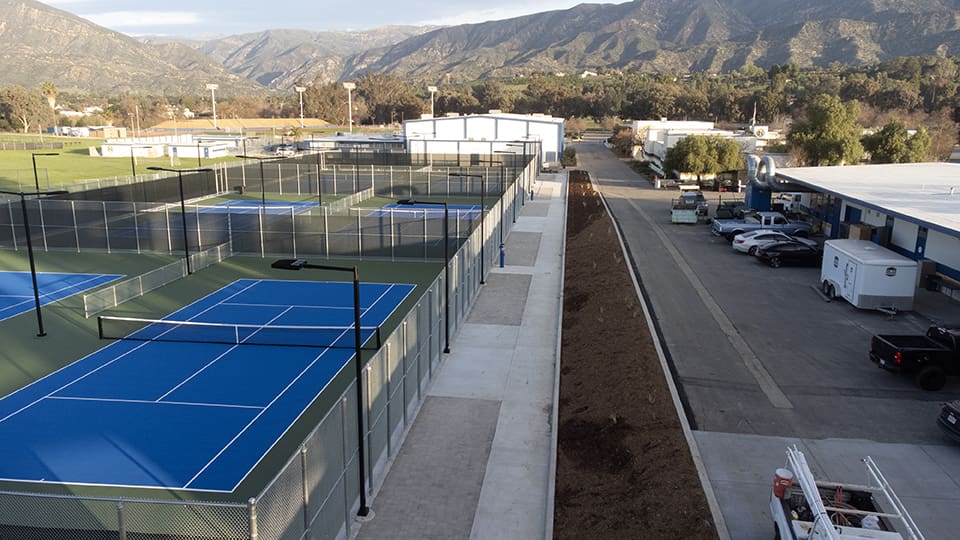 A high-angle view of multiple outdoor tennis courts with blue and green surfaces, adjacent to a parking area and buildings, set against a backdrop of mountains and trees.