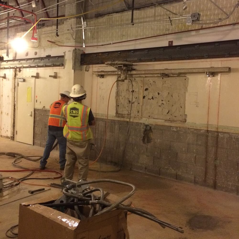 Two construction workers in safety gear and hard hats are working on a wall in an industrial space. One operates a drill, while the other observes. The floor is cluttered with tools and materials.