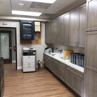 A modern office supply room with wooden cabinets, a printer, organized files, and boxes on the counter and floor. The room has wooden flooring and a door leading out.