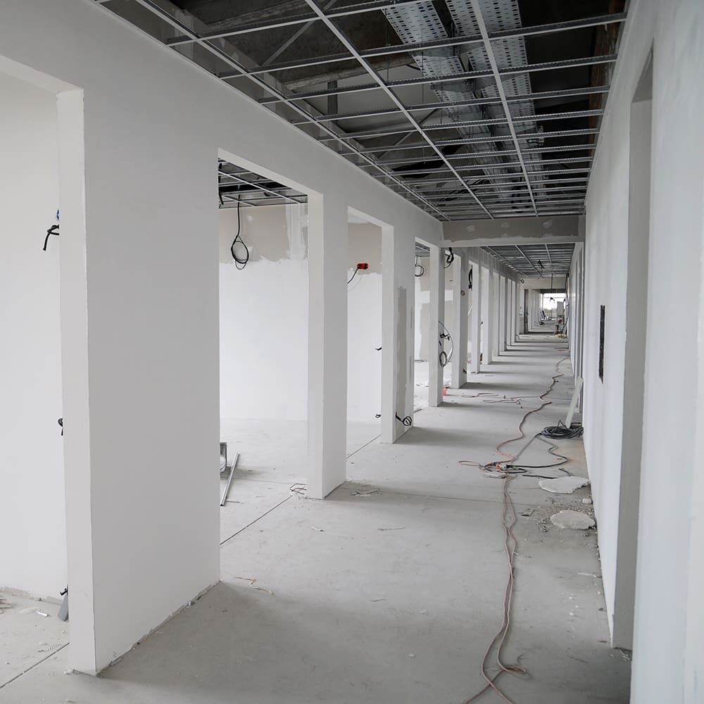 A long corridor in a building under construction, with exposed ceiling panels, bare walls, and construction materials scattered on the floor.