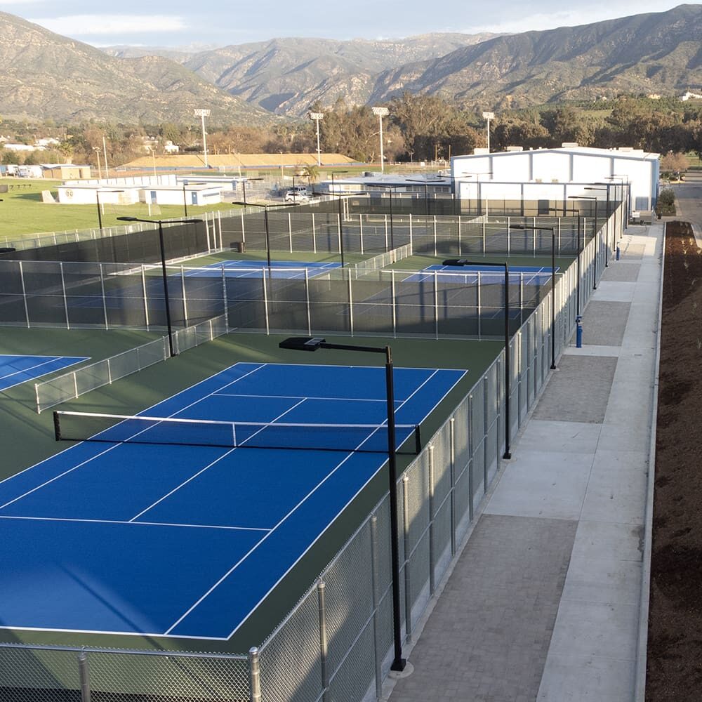 View of multiple tennis courts with blue and green surfaces, surrounded by chain-link fencing, situated near mountains and adjacent to a large building.